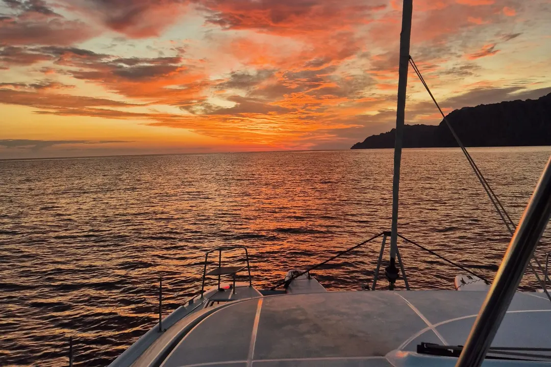 Sonnenaufgang vom Vordeck: der Himmel in Orange, das Meer glänzt, rechts eine Felsküste